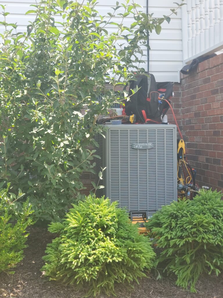 Outdoor heat pump unit installed beside a brick home surrounded by green shrubs