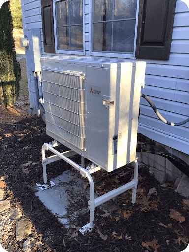 Mitsubishi outdoor heat pump condenser unit mounted on a metal stand beside a blue-sided home, with bare trees in the background