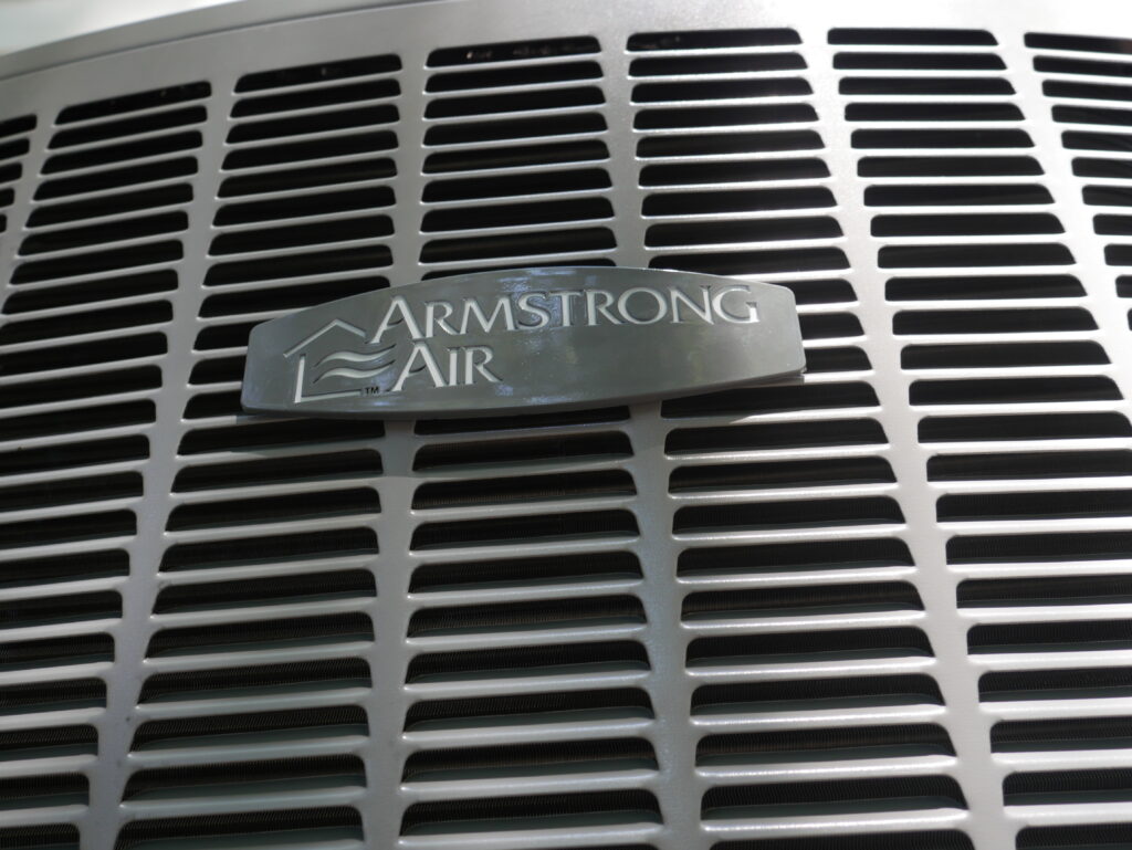 Close-up angled view of the Armstrong Air badge on the metal grille of an outdoor heat pump condenser unit